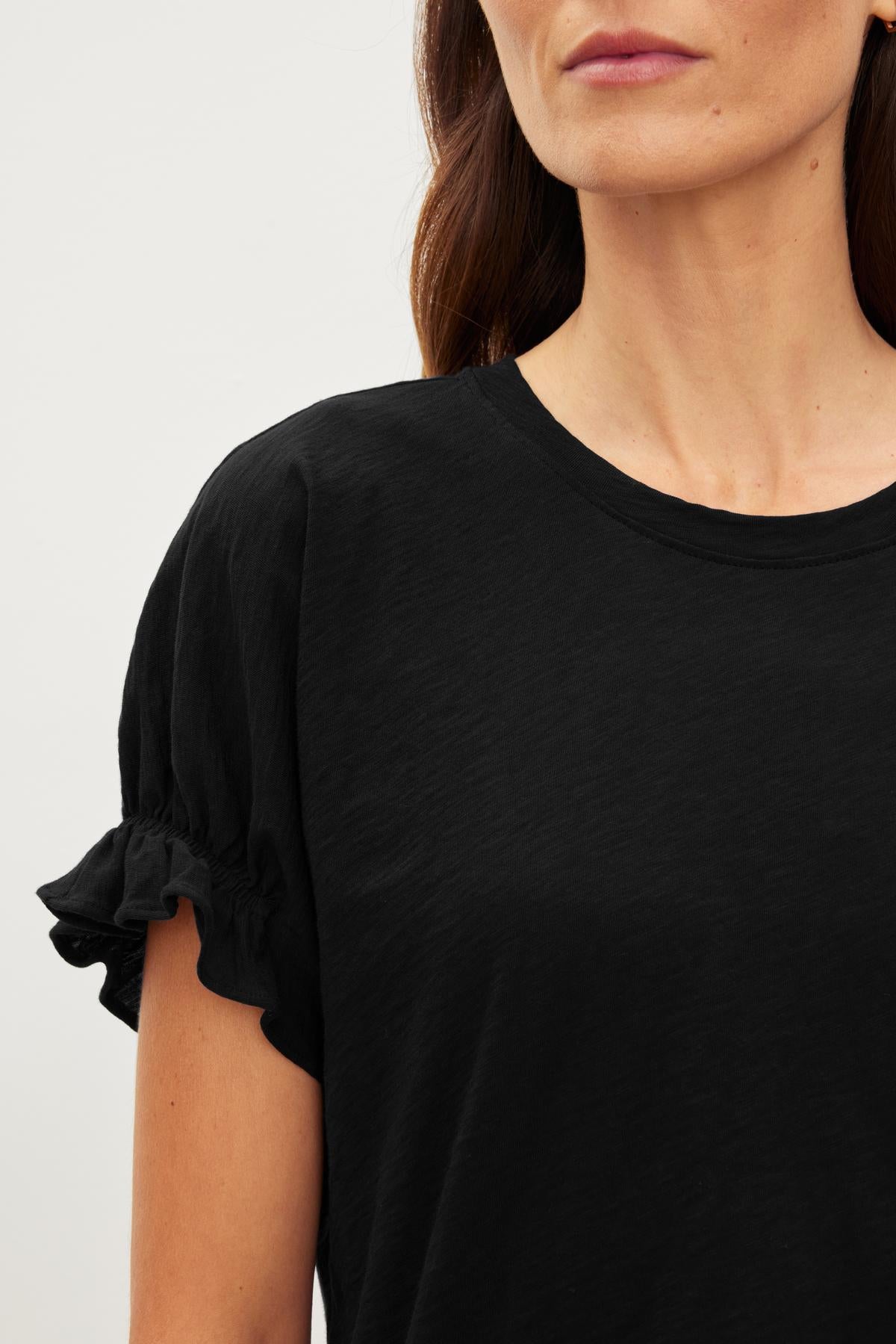   Close-up of a person wearing a black, short-sleeved MIMI CREW NECK TEE by Velvet by Graham & Spencer with ruffle details near the sleeves. The relaxed fit cotton slub fabric adds to its casual charm. The shot is cropped at the shoulders and does not include the full face. 