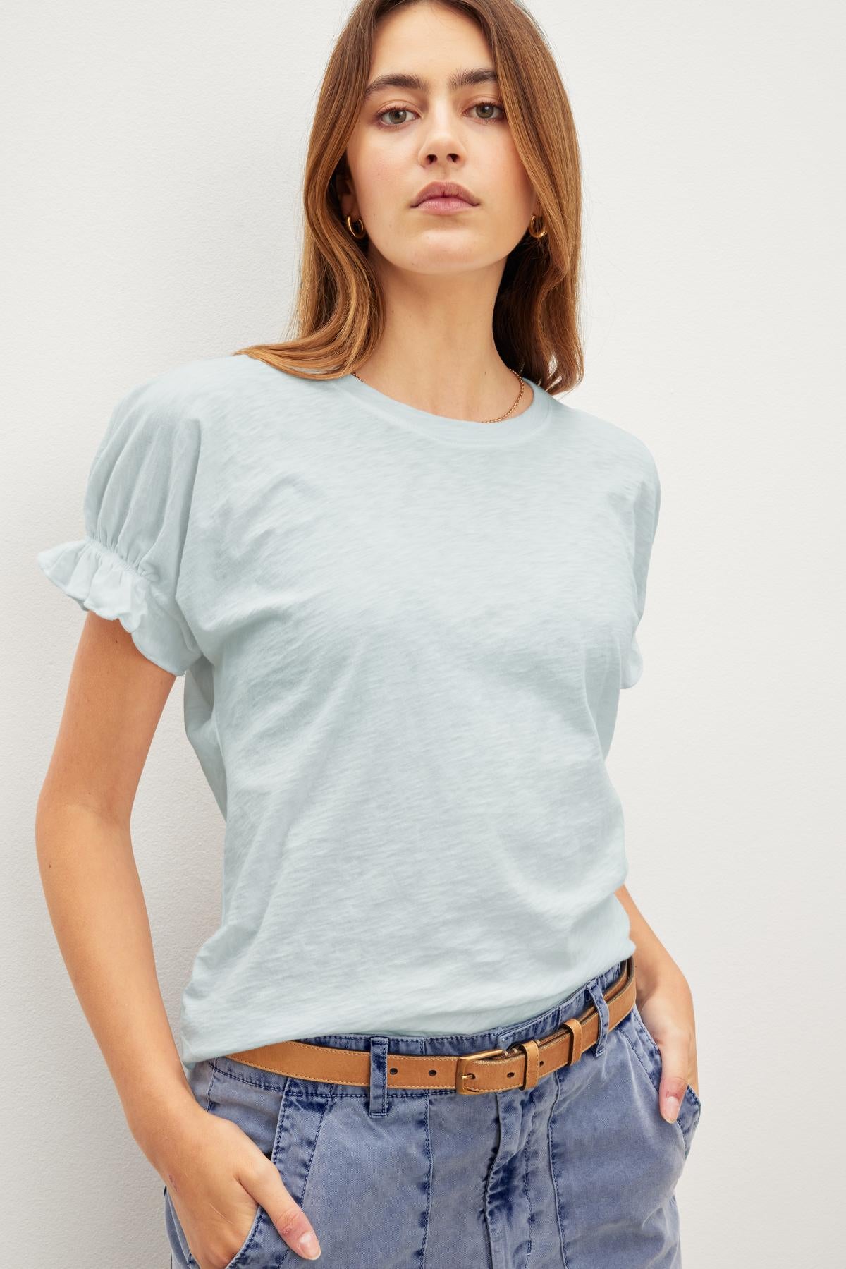   A woman stands against a plain background, wearing the MIMI CREW NECK TEE by Velvet by Graham & Spencer and light blue pants. Her hands are in her pockets, and she has a neutral expression, showcasing the relaxed fit. 