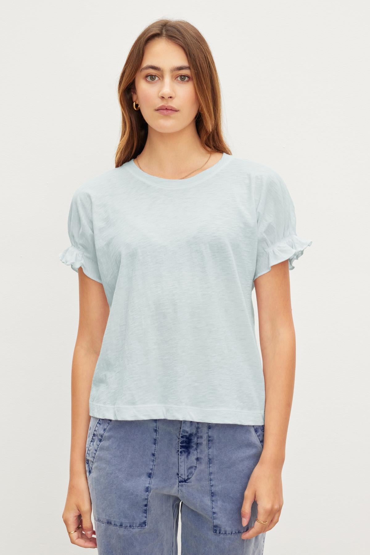   A person stands against a plain background, wearing the MIMI CREW NECK TEE by Velvet by Graham & Spencer with ruffle detail and blue jeans. The relaxed fit of the cotton slub fabric adds to the effortless chic style. 