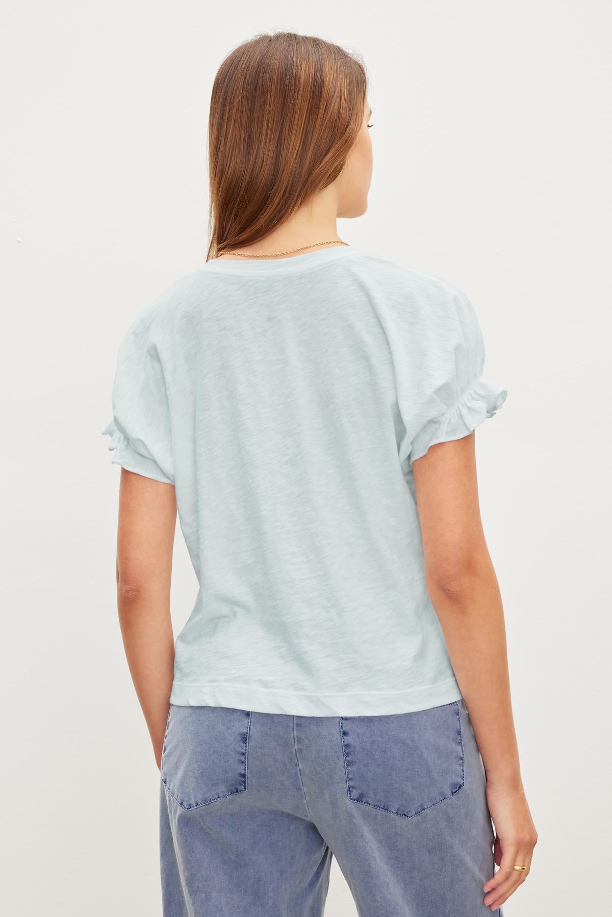   A person with long hair is standing and facing away, wearing a light blue MIMI CREW NECK TEE by Velvet by Graham & Spencer with ruffled cuffs and light blue relaxed fit pants. 