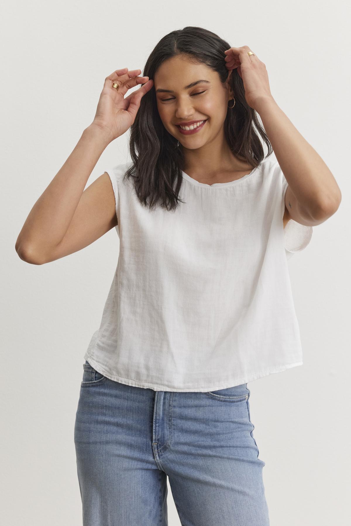A person with long dark hair smiles while adjusting their hair, wearing a white sleeveless top and blue jeans against a plain white background. The casual basic outfit features the DANIELLA CREW NECK TEE by Velvet by Graham & Spencer, made from a lightweight cotton woven fabric that's perfect for everyday wear.-37074420891841