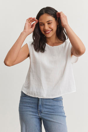 A person with long dark hair smiles while adjusting their hair, wearing a white sleeveless top and blue jeans against a plain white background. The casual basic outfit features the DANIELLA CREW NECK TEE by Velvet by Graham & Spencer, made from a lightweight cotton woven fabric that's perfect for everyday wear.