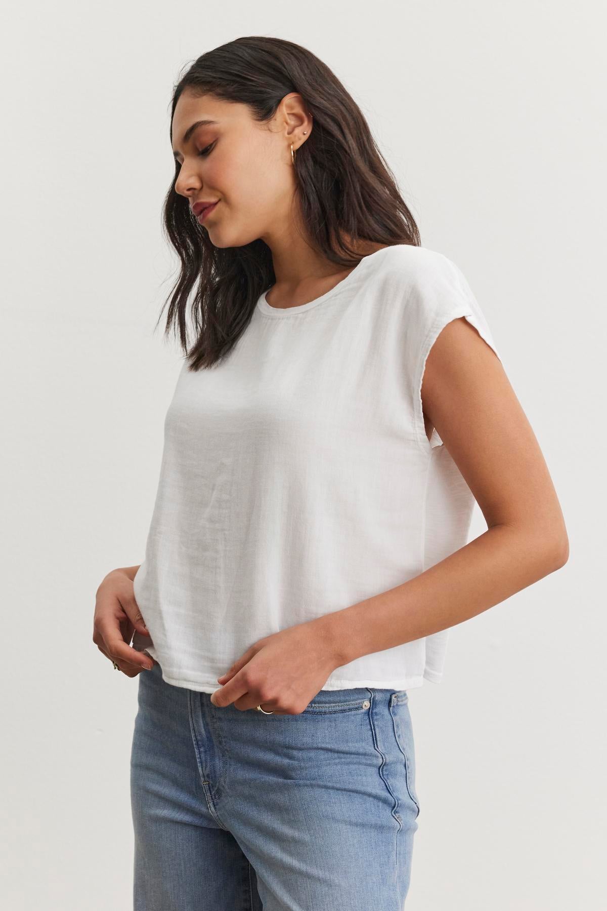   A woman with long dark hair wearing a white DANIELLA CREW NECK TEE by Velvet by Graham & Spencer and blue jeans, showcasing a casual basic look, stands against a plain, light-colored background. The lightweight cotton woven fabric of her cropped silhouette shirt complements her style effortlessly. 