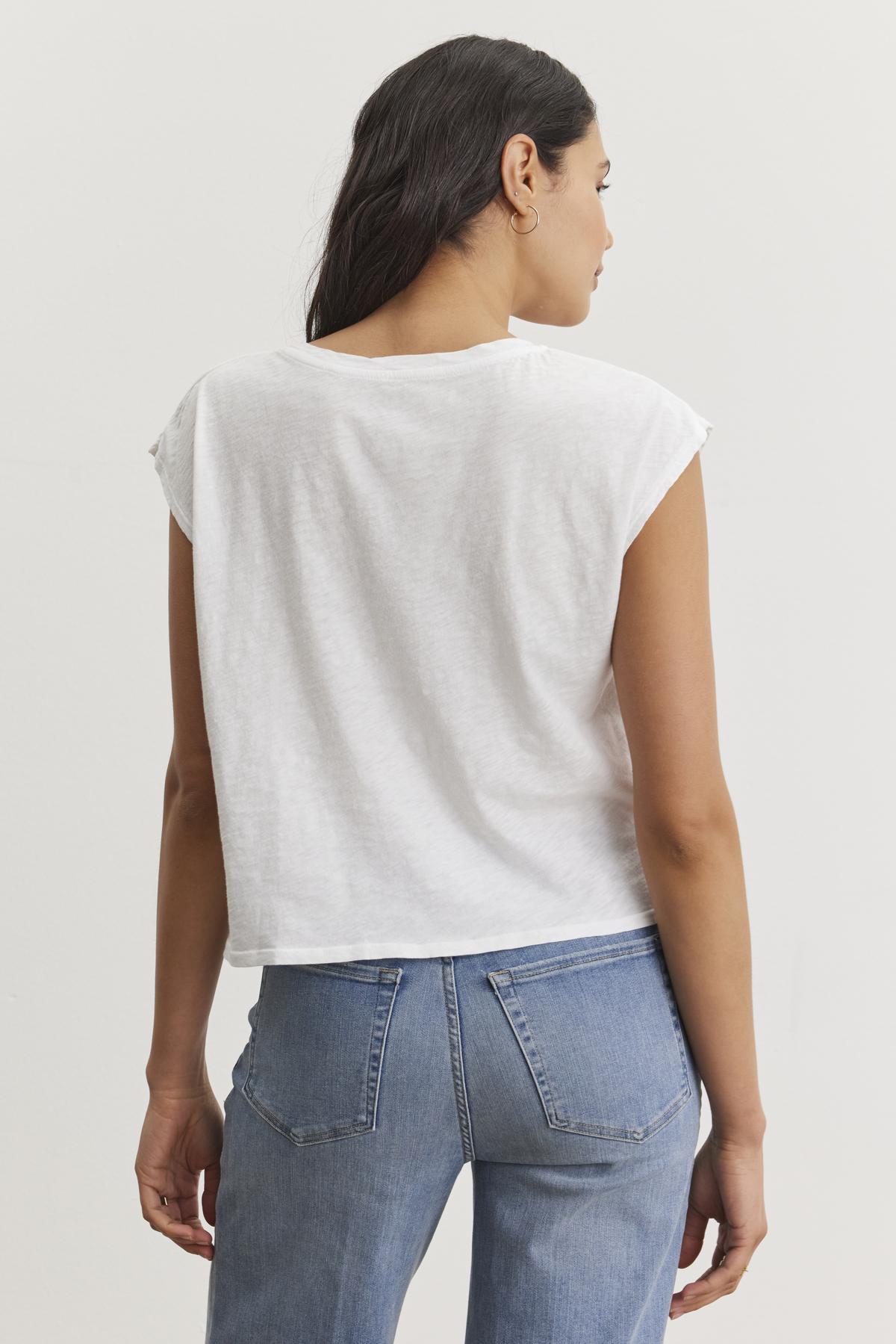 A woman with long brown hair is seen from the back, wearing a white DANIELLA CREW NECK TEE made of lightweight cotton woven fabric and light blue jeans by Velvet by Graham & Spencer. She stands against a plain white background, embodying a casual basic style.-37074420957377