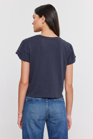 Woman viewed from behind, wearing a dark blue cotton slub DELLA TEE with pleated sleeve detail and blue jeans, standing against a white background. (Brand Name: Velvet by Graham & Spencer)