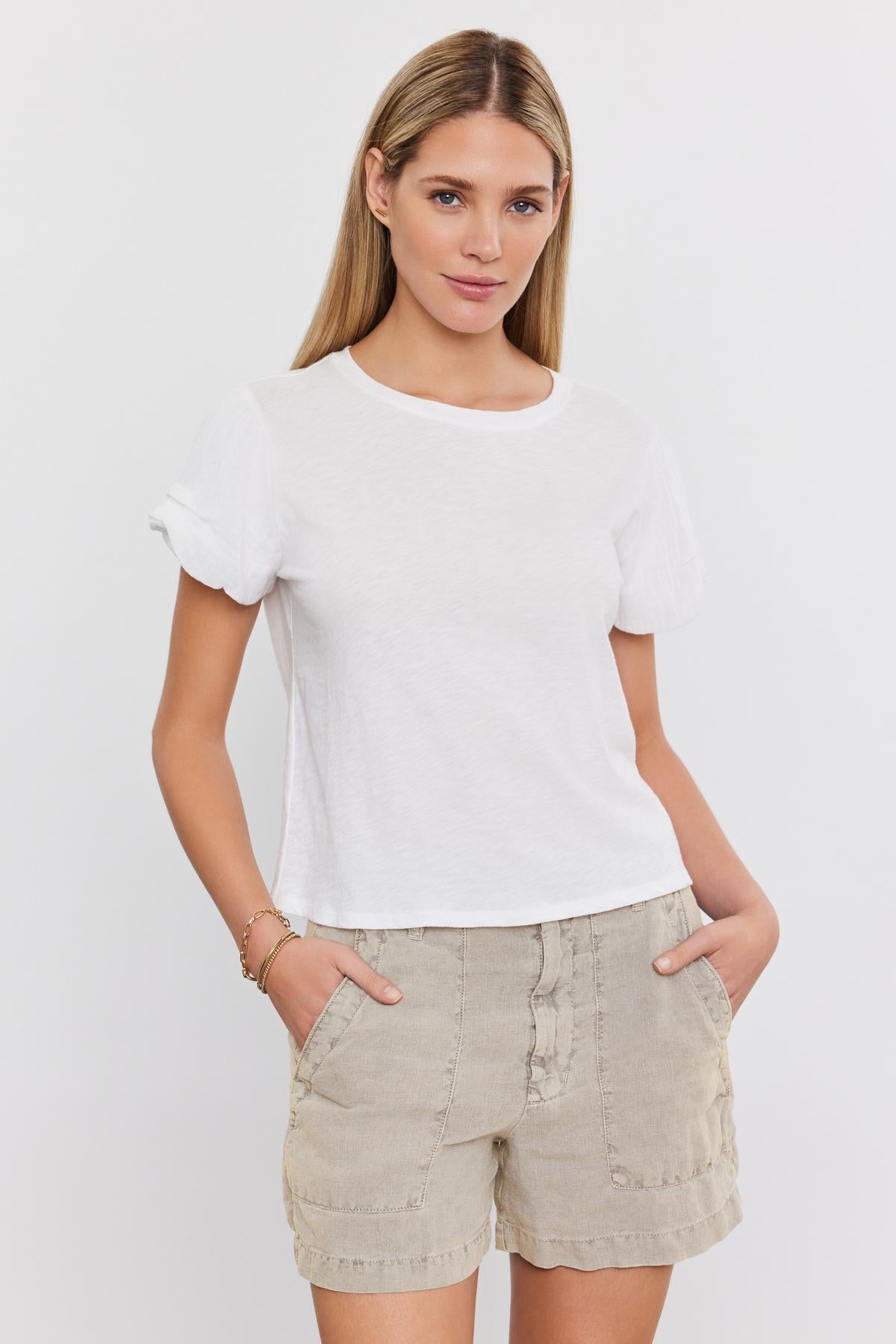   A woman with long hair, wearing a white t-shirt and Velvet by Graham & Spencer's FALLON HEAVY LINEN SHORT, stands against a plain background with her hands in her pockets. 