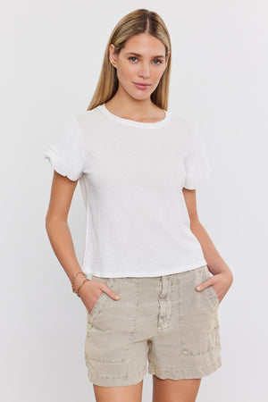 A woman with long hair, wearing a white t-shirt and Velvet by Graham & Spencer's FALLON HEAVY LINEN SHORT, stands against a plain background with her hands in her pockets.