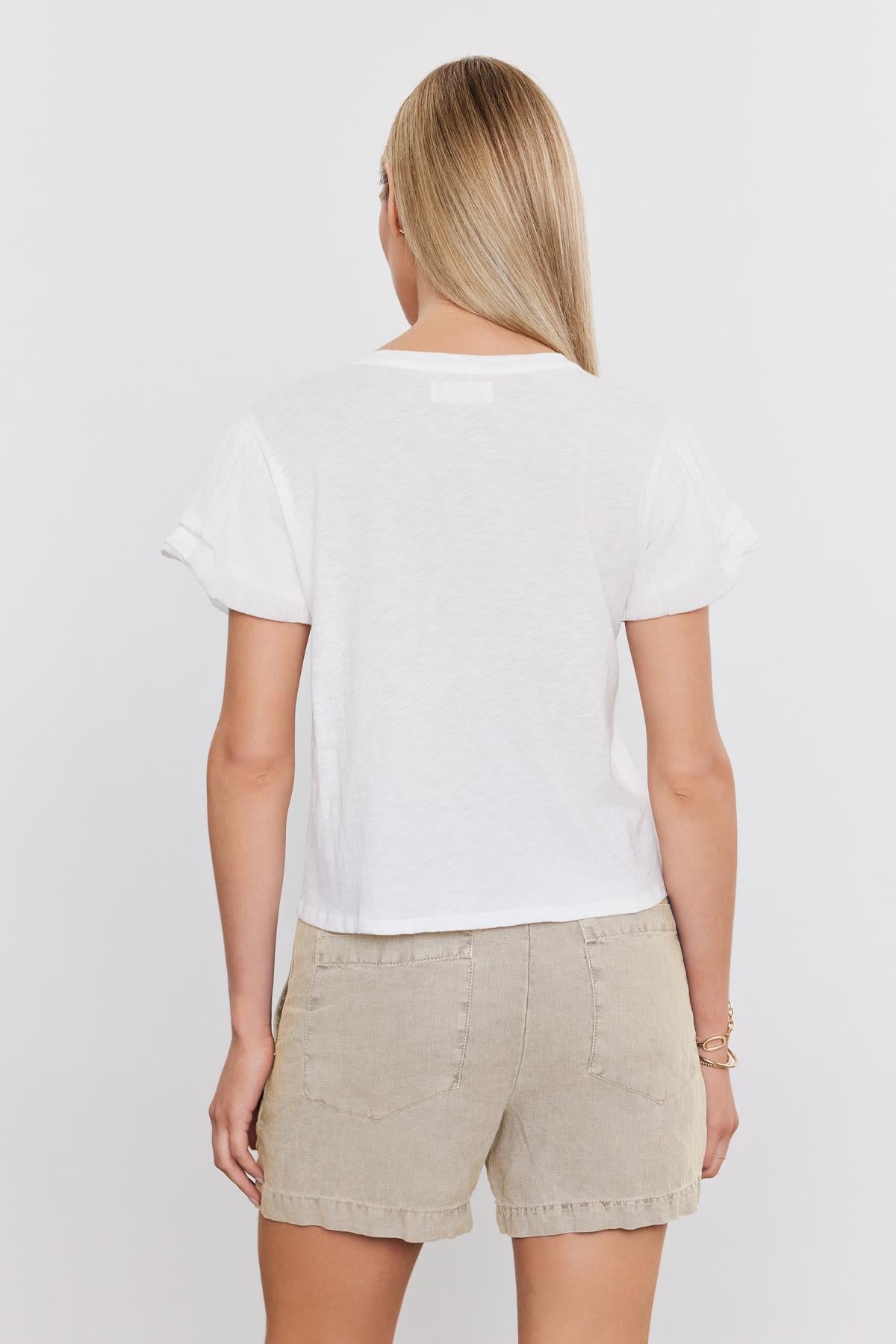   A person with long blonde hair, facing away from the camera, is wearing a white T-shirt and beige FALLON HEAVY LINEN SHORT by Velvet by Graham & Spencer. 