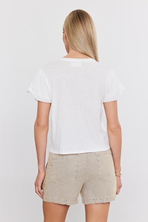 A person with long blonde hair, facing away from the camera, is wearing a white T-shirt and beige FALLON HEAVY LINEN SHORT by Velvet by Graham & Spencer.