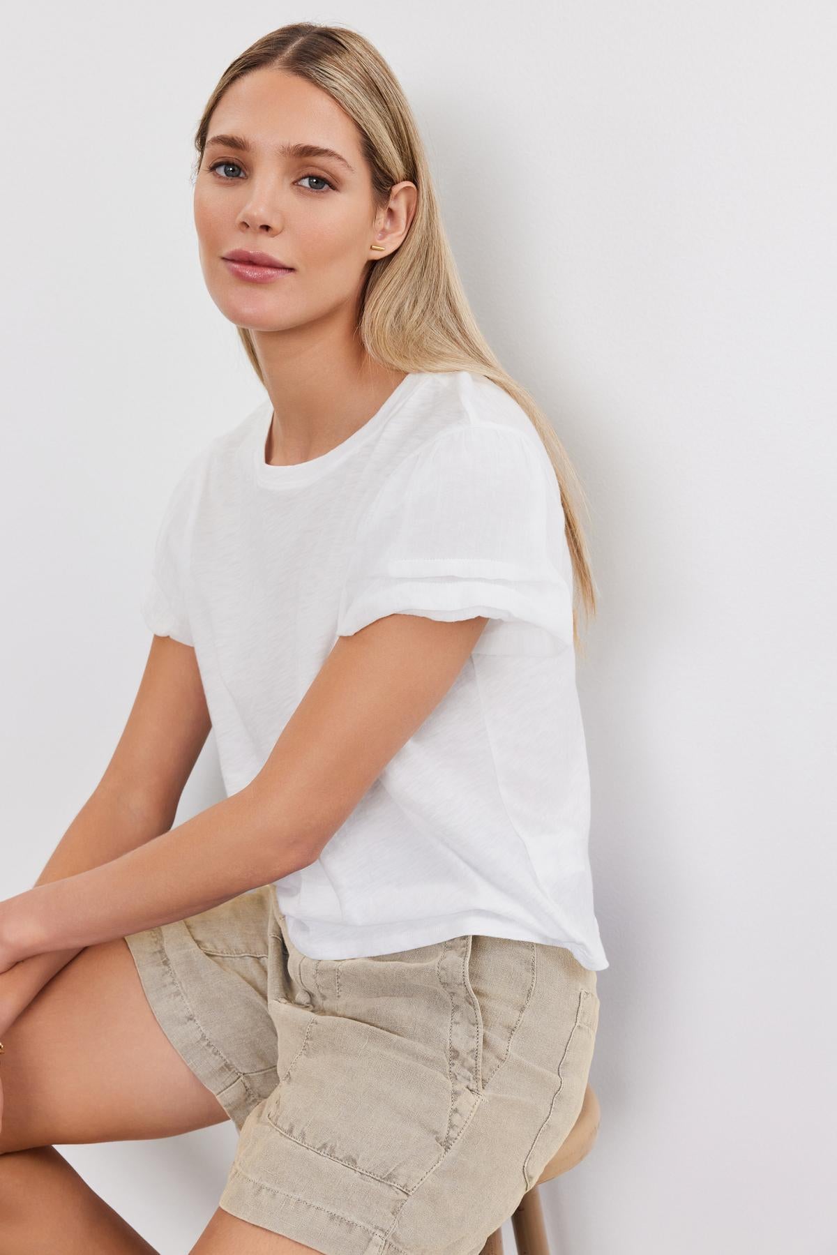   A person with long blonde hair wearing a white T-shirt and the comfortable, beige Fallon Heavy Linen Shorts from Velvet by Graham & Spencer sits on a stool against a plain white background. 