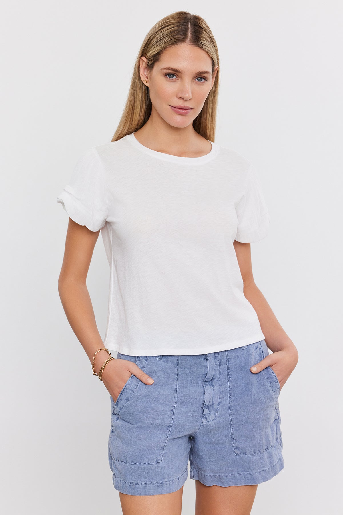   Woman with long blonde hair wearing a DELLA TEE by Velvet by Graham & Spencer and blue shorts, standing with hands in pockets, against a white background. 