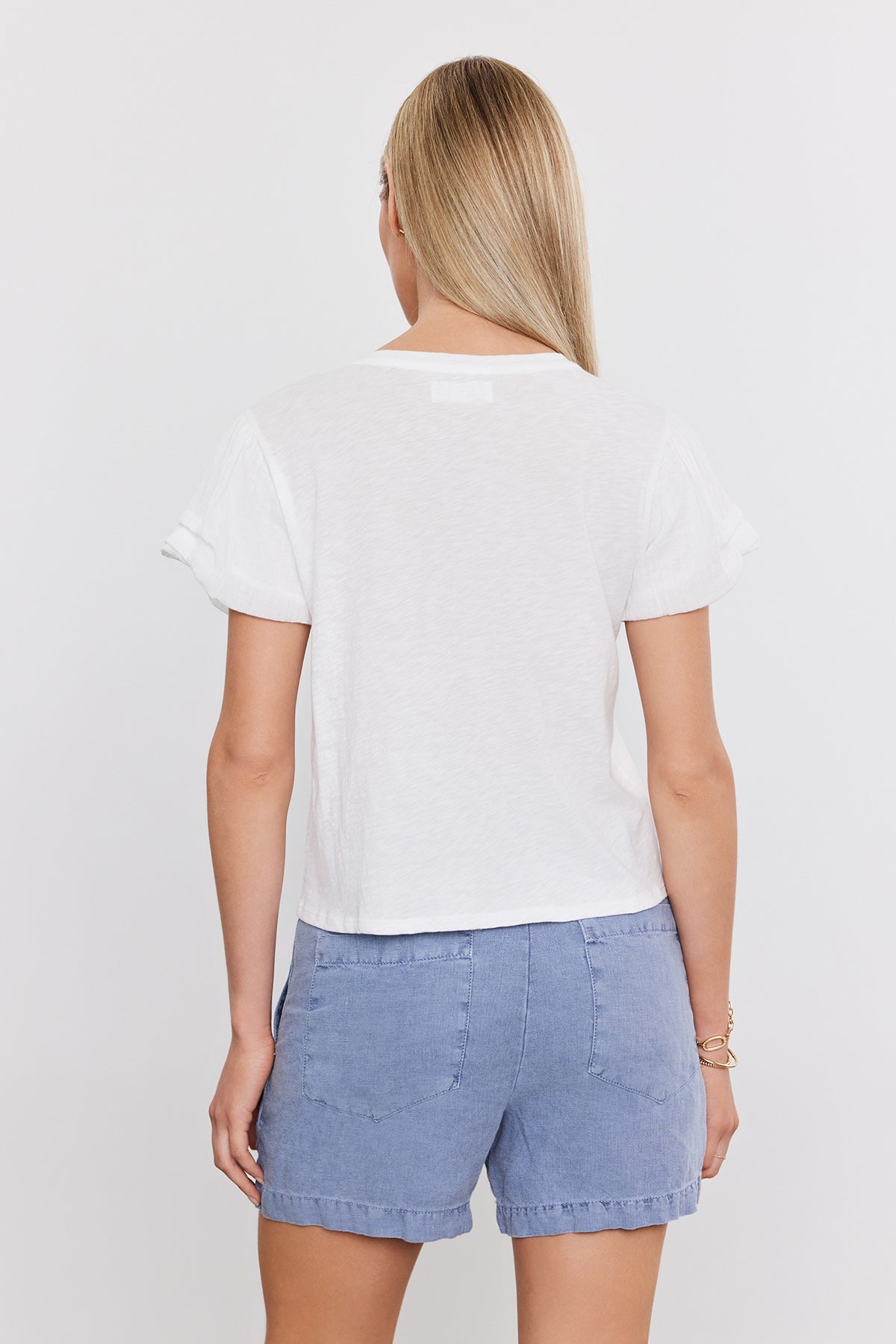   A person with long blond hair is viewed from the back, wearing a white short-sleeve DELLA TEE by Velvet by Graham & Spencer and light blue shorts. 