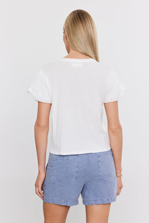 A person with long blond hair is viewed from the back, wearing a white short-sleeve DELLA TEE by Velvet by Graham & Spencer and light blue shorts.