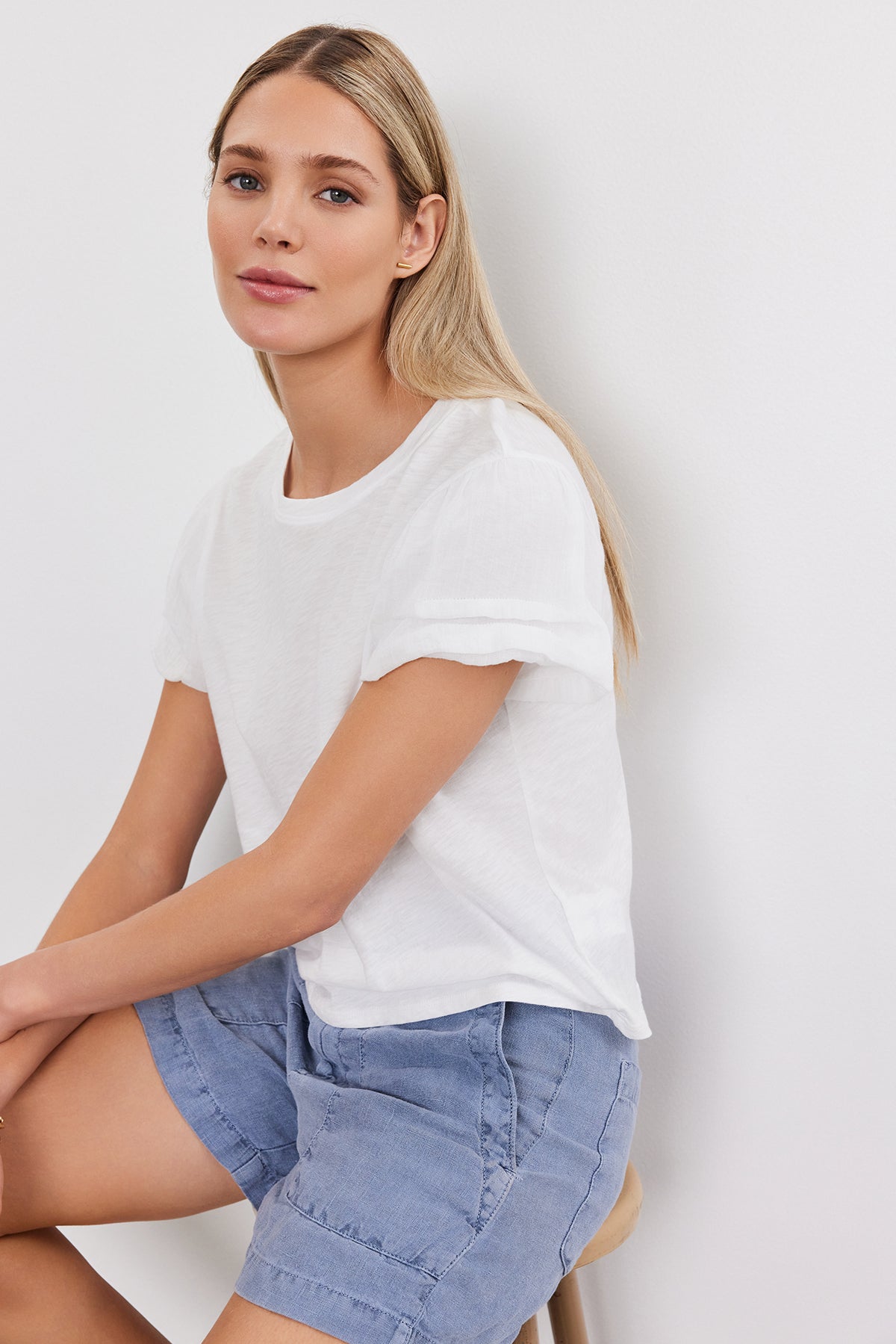   A woman with long blonde hair wearing a white DELLA TEE by Velvet by Graham & Spencer and blue shorts sits on a stool against a plain white background. 