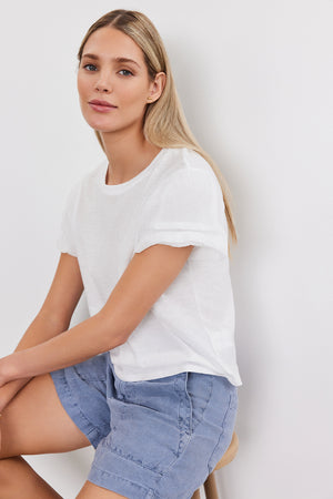 A woman with long blonde hair wearing a white DELLA TEE by Velvet by Graham & Spencer and blue shorts sits on a stool against a plain white background.