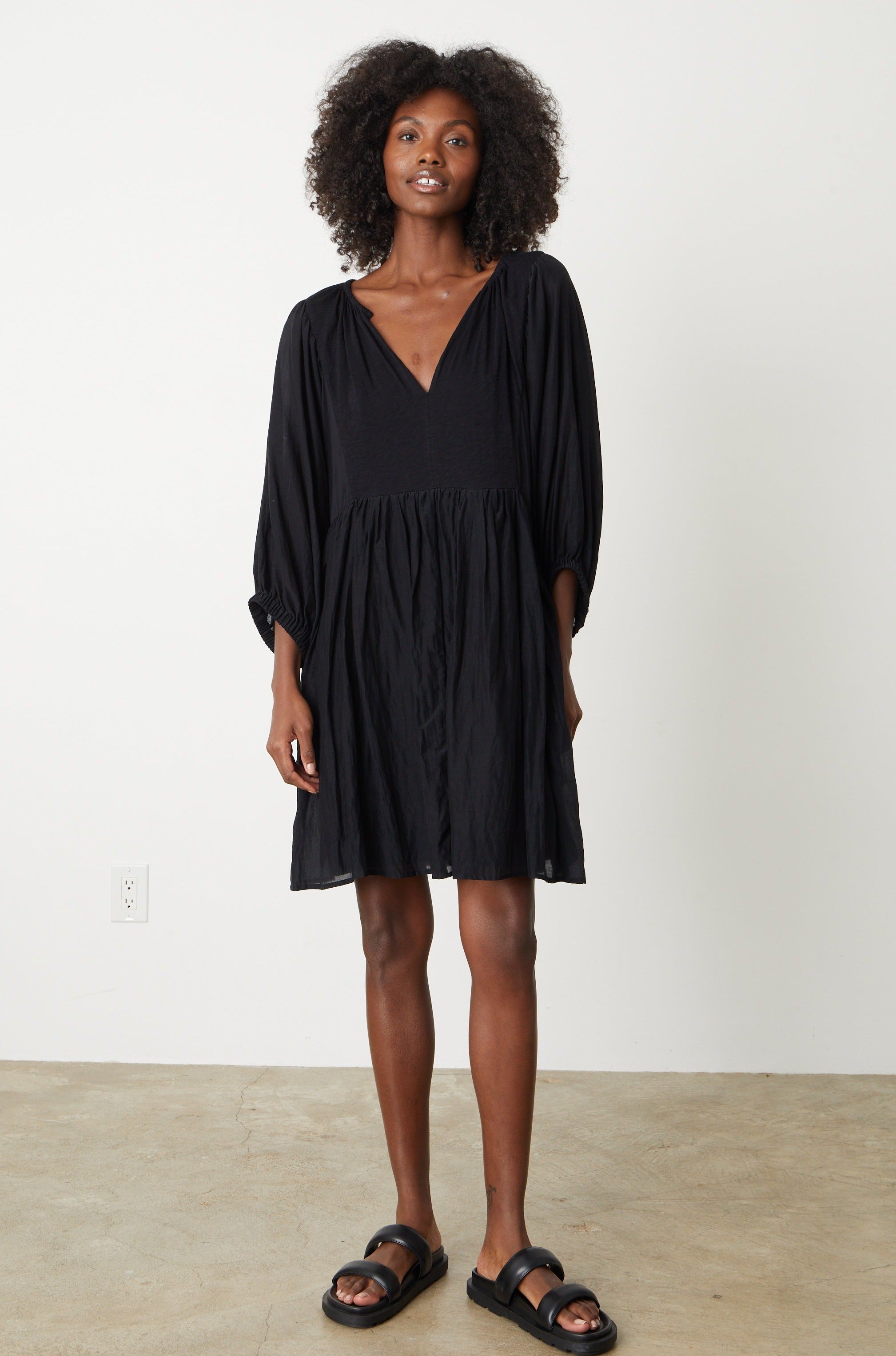   A woman wearing a Velvet by Graham & Spencer ERIN SHIRRED WAIST DRESS in black and sandals. 