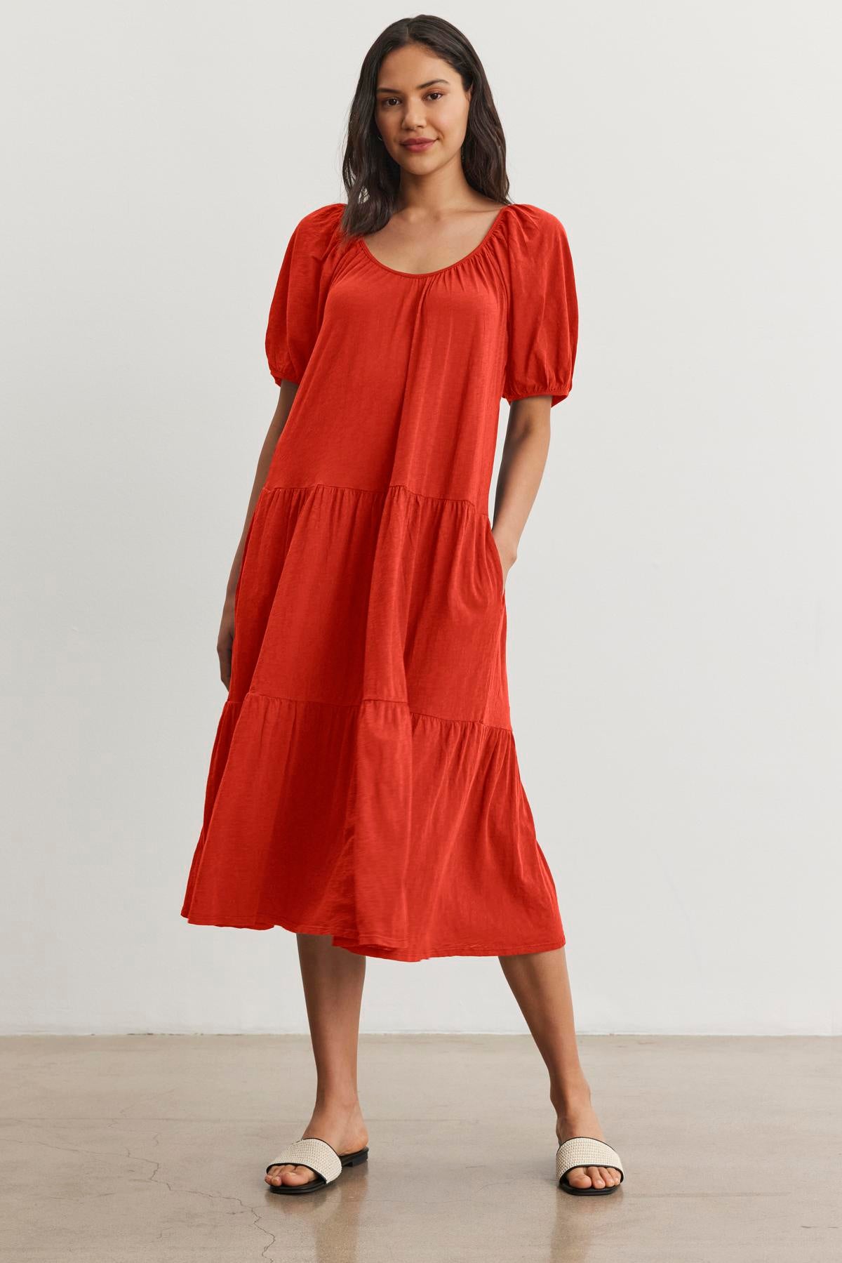   A woman stands against a plain backdrop wearing the Velvet by Graham & Spencer JANE DRESS, a red, short-sleeve, tiered midi dress with a drop-waist silhouette, and white sandals, perfect for summer vacations. 