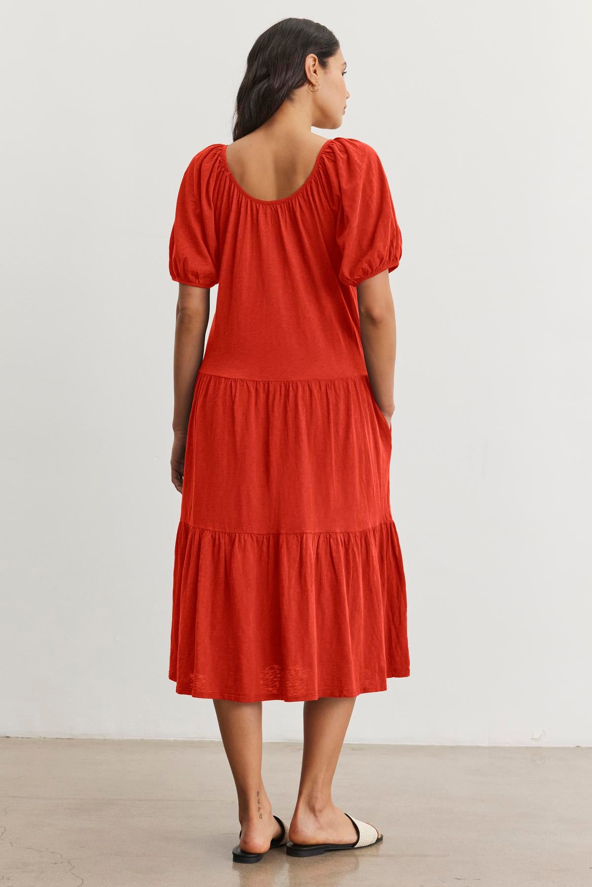   A woman wearing the Velvet by Graham & Spencer JANE DRESS, a knee-length red dress with short puffed sleeves and a scooped neckline, stands facing away. The lightweight cotton slub knit fabric adds to the casual vibe, perfect for summer vacations. She is barefoot on a plain, light-colored floor against a white wall. 