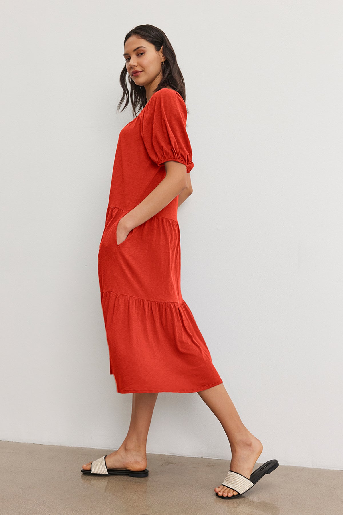 A woman stands against a plain white wall, wearing the JANE DRESS by Velvet by Graham & Spencer and black-and-white sandals—a perfect drop-waist silhouette for summer vacations.-37452667224257