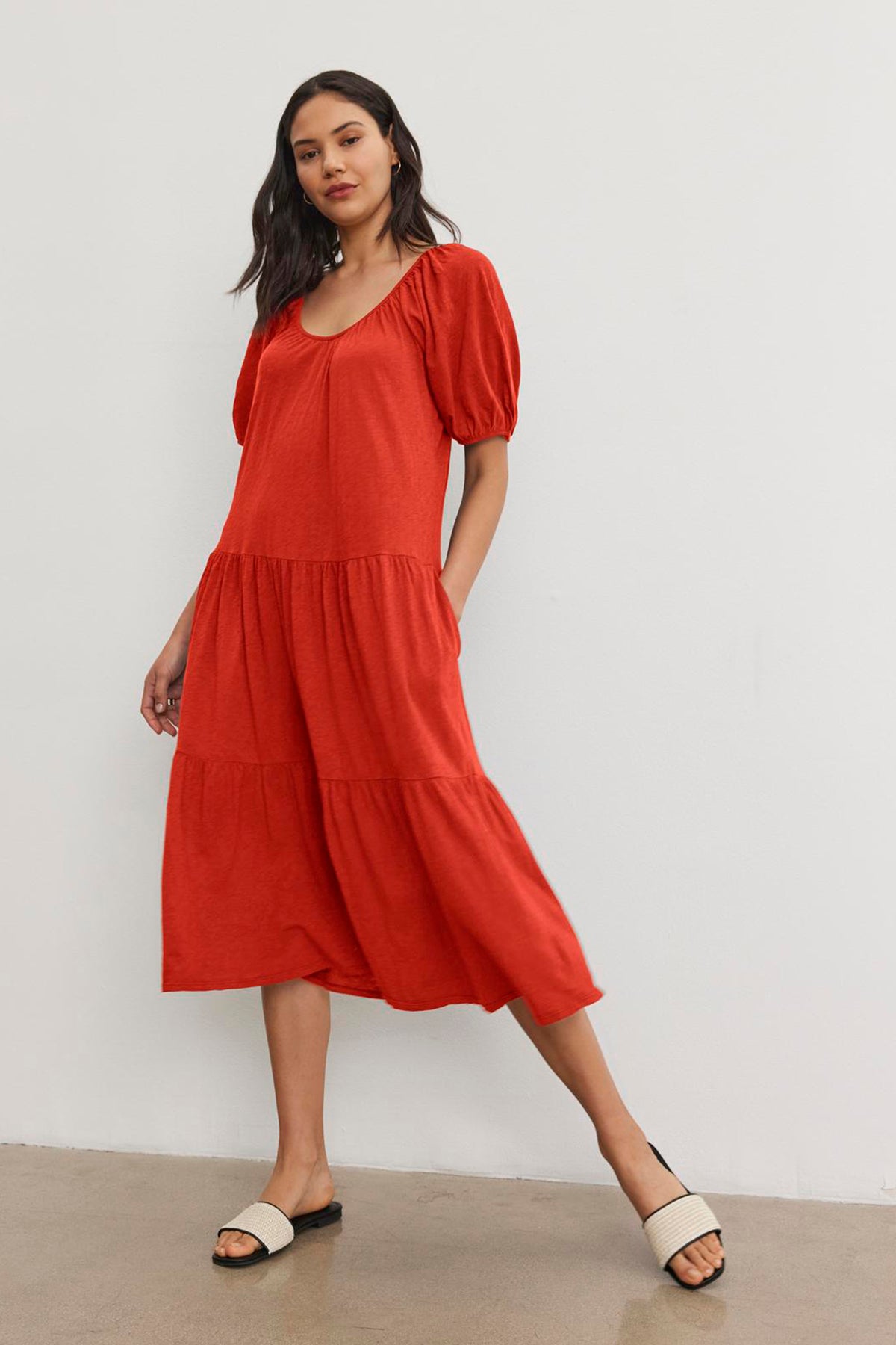   A woman in a red, short-sleeved, JANE DRESS by Velvet by Graham & Spencer with a drop-waist silhouette stands against a plain white wall, wearing black and white open-toed sandals. 