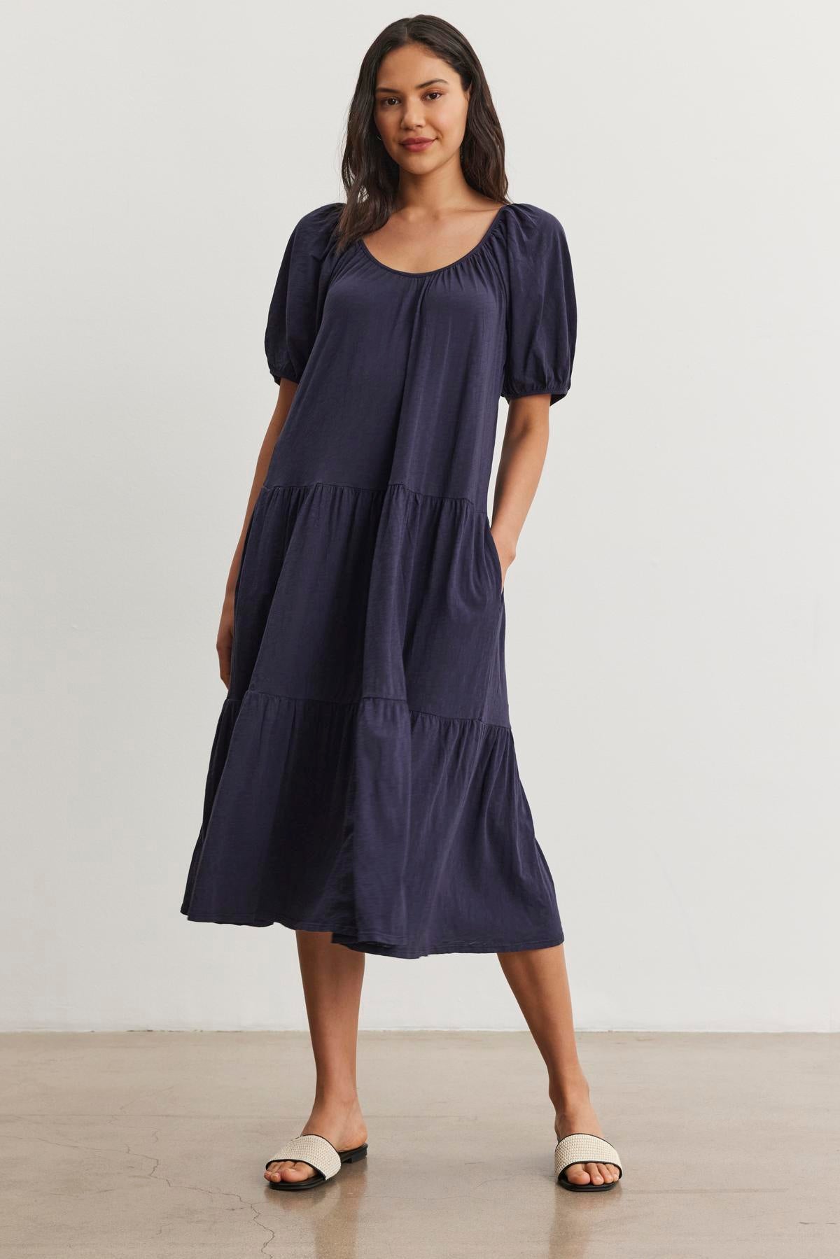   A woman wearing a navy blue, short-sleeved, knee-length JANE DRESS by Velvet by Graham & Spencer with a drop-waist silhouette stands against a plain white background. She is also wearing white sandals. 