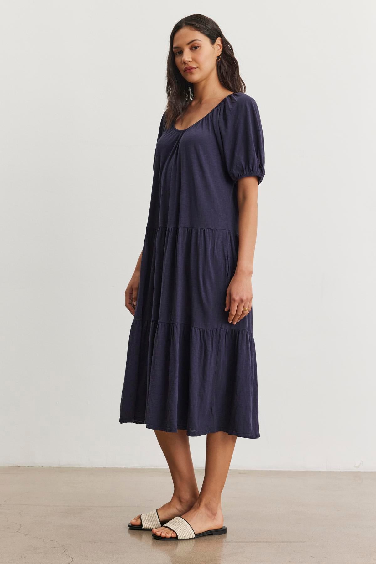   A person is standing and wearing a navy blue, mid-length JANE DRESS by Velvet by Graham & Spencer with puffed sleeves and sandals. The cotton slub knit fabric gives it a relaxed feel, perfect for summer vacations. The background is plain white. 