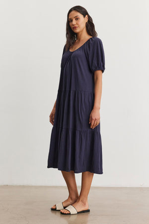 A person is standing and wearing a navy blue, mid-length JANE DRESS by Velvet by Graham & Spencer with puffed sleeves and sandals. The cotton slub knit fabric gives it a relaxed feel, perfect for summer vacations. The background is plain white.