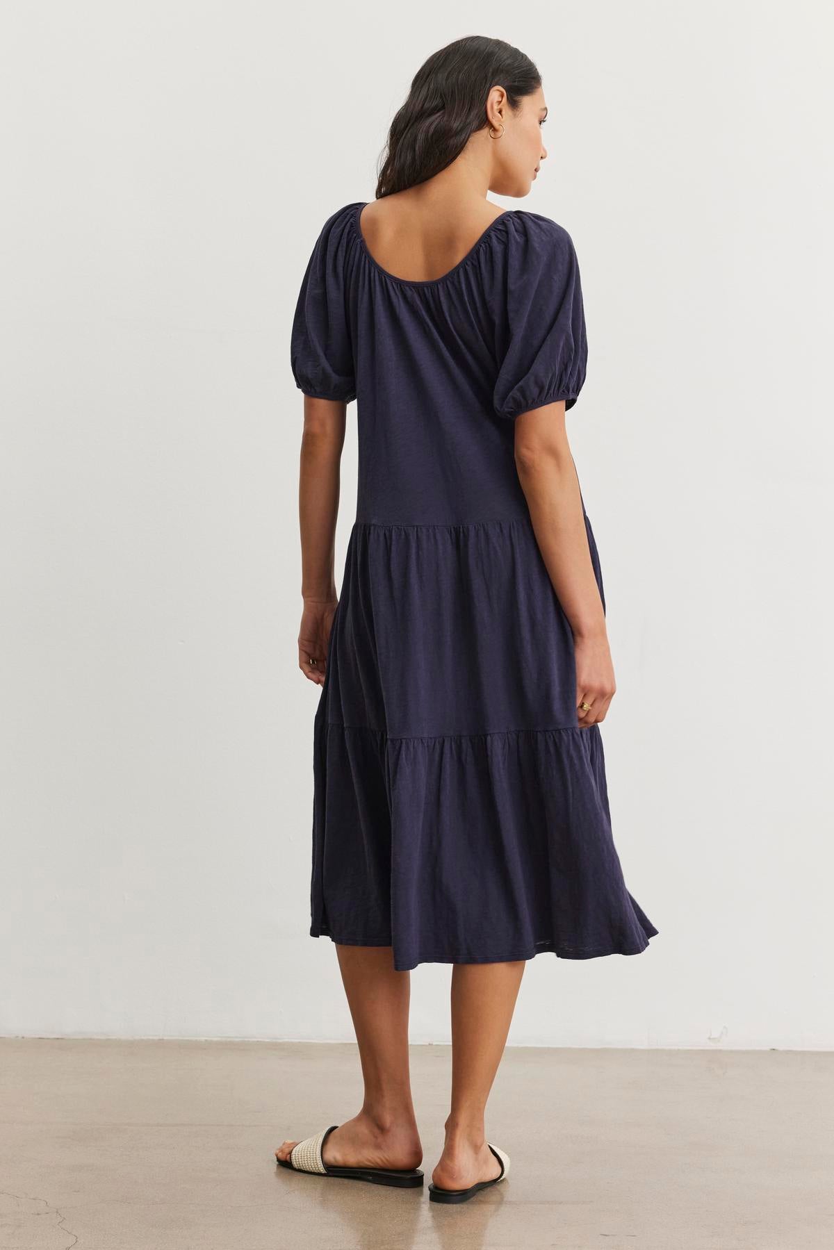   A person wearing a navy blue, tiered midi dress with short puff sleeves and a drop-waist silhouette stands facing away, showcasing the back of the JANE DRESS by Velvet by Graham & Spencer. They are also wearing flat, open-toed sandals. Perfect for summer vacations, this outfit combines style and comfort effortlessly. 