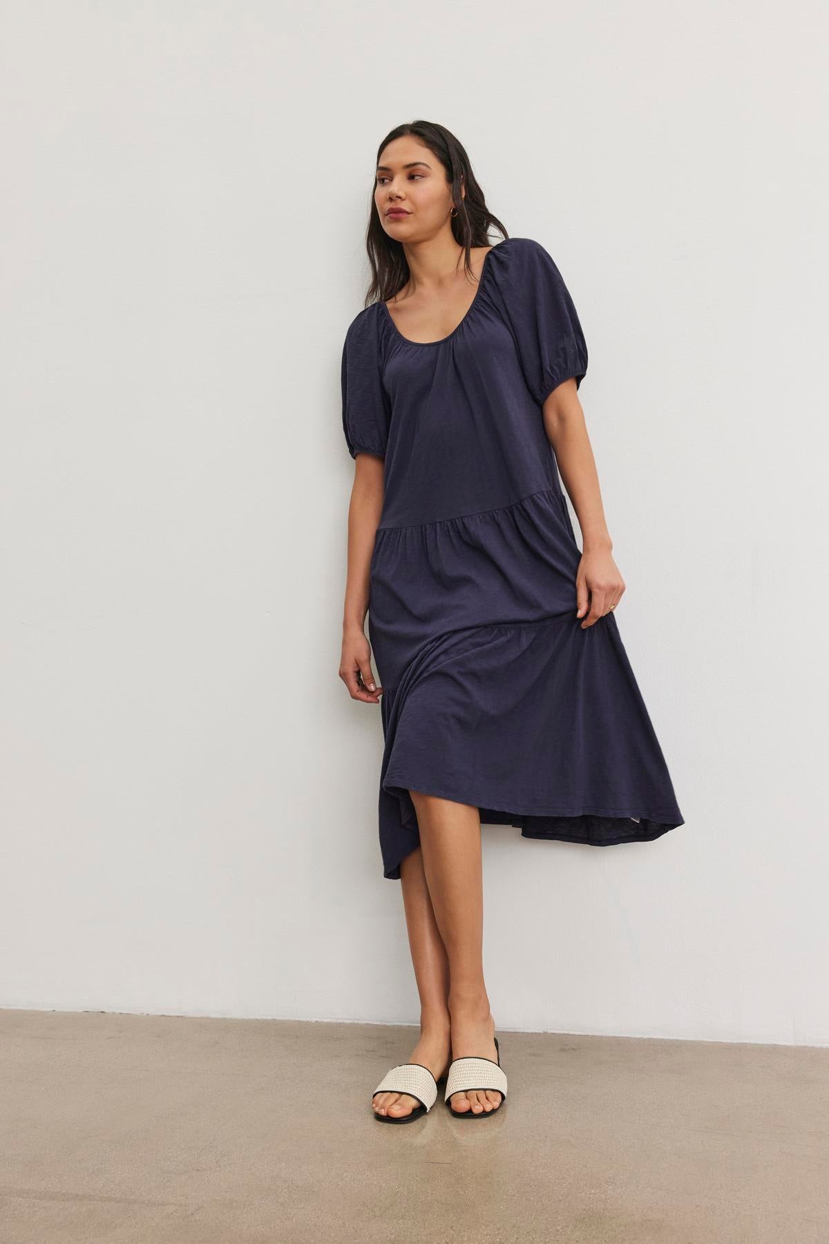   A woman stands against a plain white wall, wearing the JANE DRESS by Velvet by Graham & Spencer, a dark blue knee-length dress with short puffy sleeves and a flattering drop-waist silhouette, paired with white open-toe sandals. 