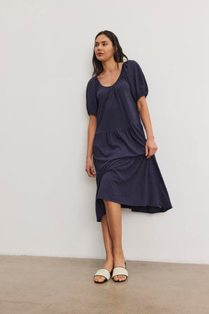 A woman stands against a plain white wall, wearing the JANE DRESS by Velvet by Graham & Spencer, a dark blue knee-length dress with short puffy sleeves and a flattering drop-waist silhouette, paired with white open-toe sandals.