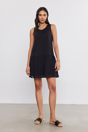 A woman in a sleeveless black Velvet by Graham & Spencer MINA DRESS with a tiered skirt and black sandals stands on a plain white background.