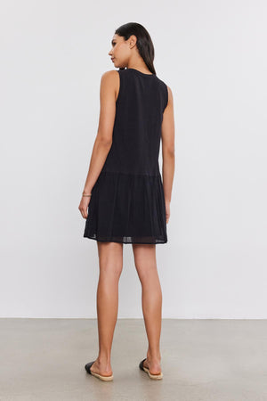 A woman wearing a sleeveless black MINA DRESS by Velvet by Graham & Spencer with a tiered skirt stands with her back to the camera, in a simple, well-lit studio setting.