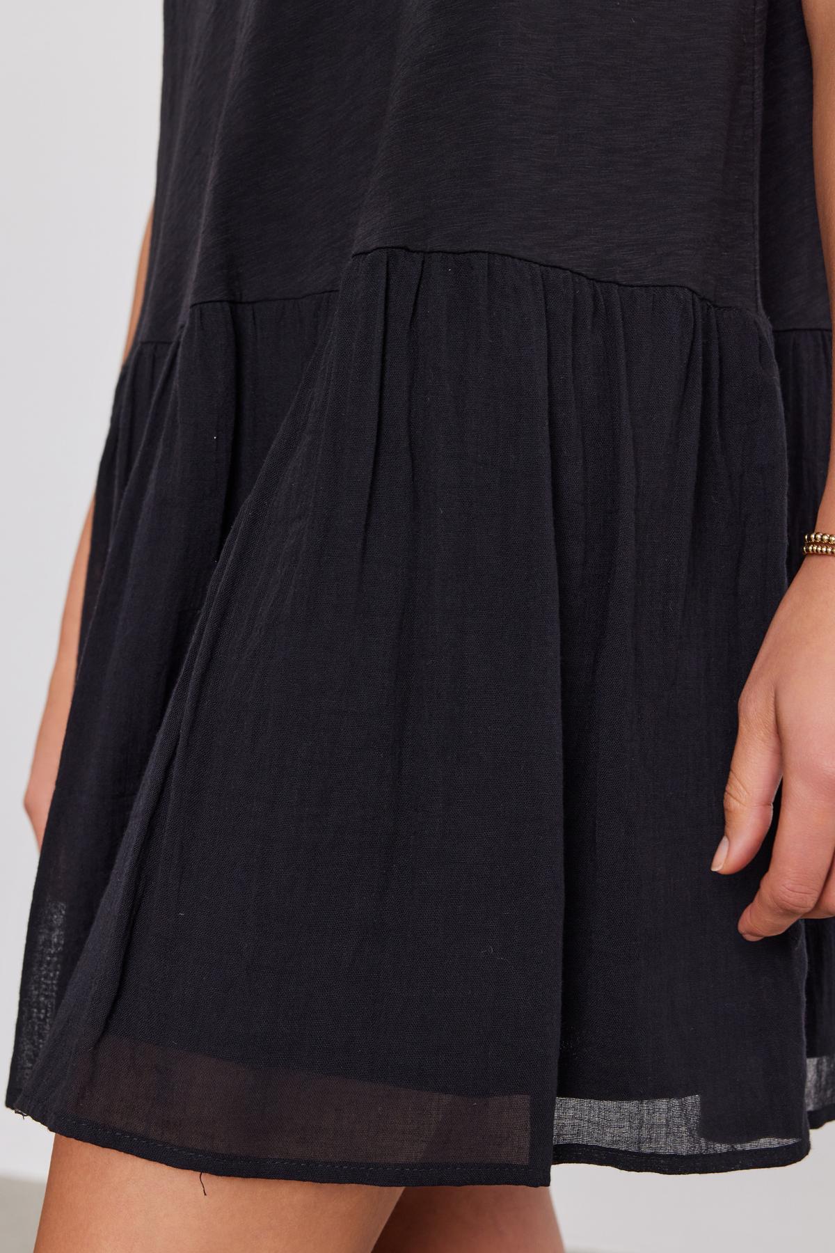 Close-up of a section of a black sleeveless MINA DRESS with a tiered, gathered skirt made from cotton slub by Velvet by Graham & Spencer, and a woman's arm visible to the right.-37054552506561