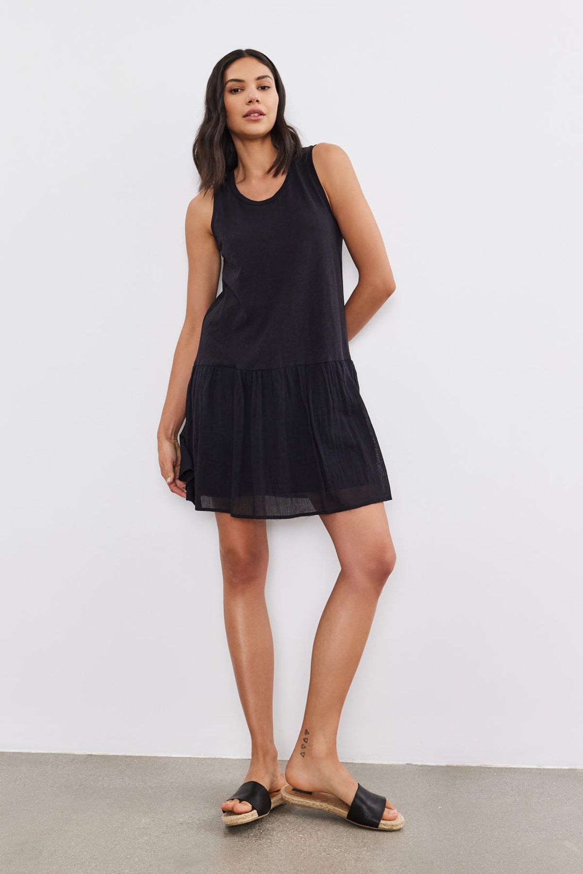 A woman with dark hair is wearing a sleeveless, black, drop-waist MINA DRESS by Velvet by Graham & Spencer featuring a tiered skirt and black slide sandals, standing against a plain white background. She has a relaxed posture with one leg slightly bent.-37054552473793