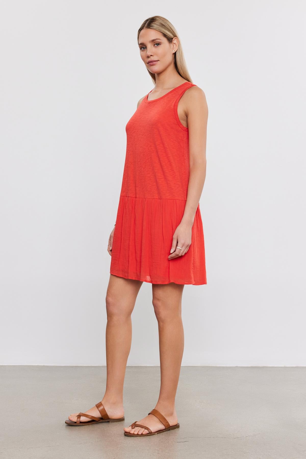 Woman standing in a studio, wearing a red sleeveless MINA DRESS with a tiered skirt and brown sandals, facing slightly right with a neutral expression by Velvet by Graham & Spencer.-36910021443777