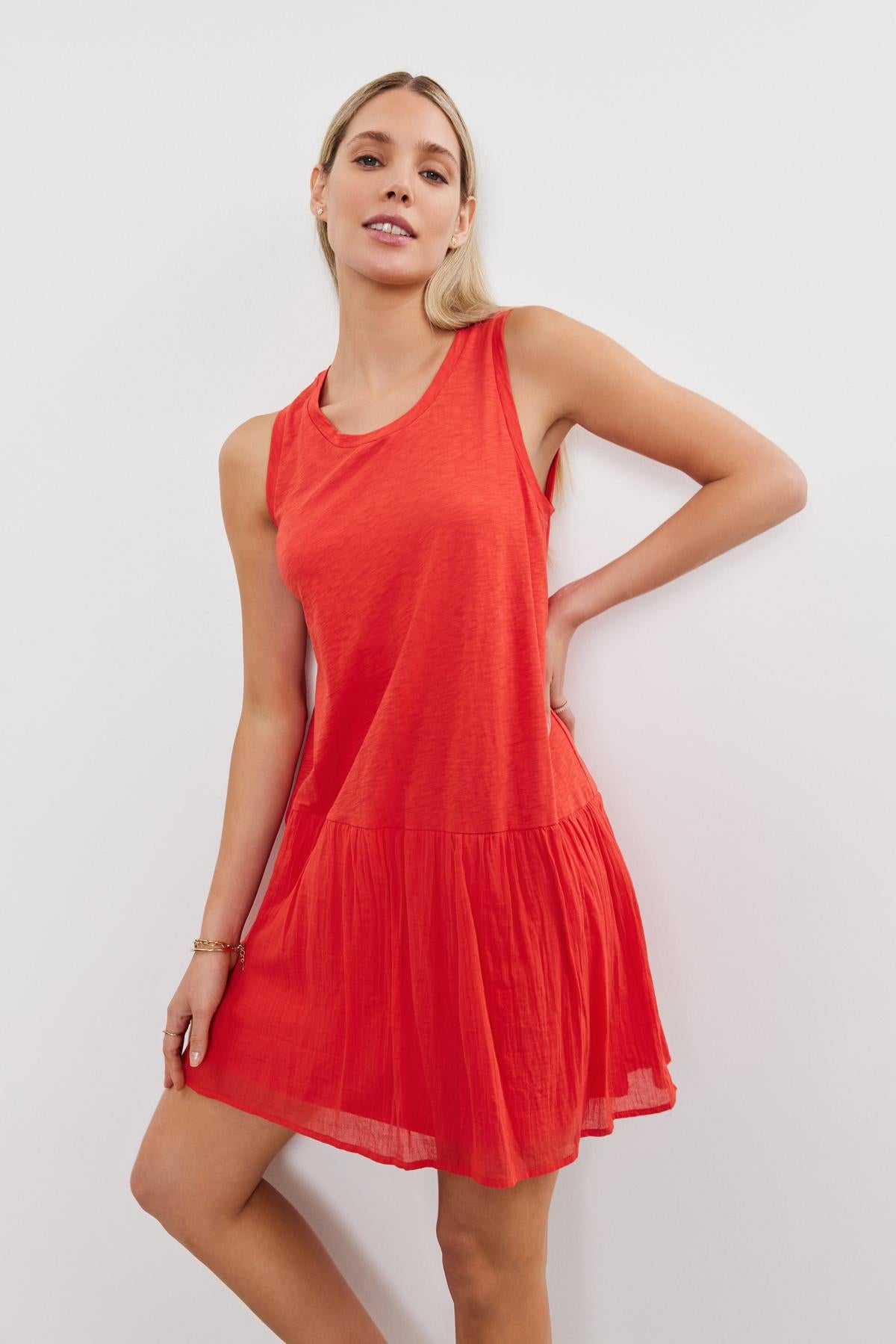 A young woman stands against a white background, wearing a casual sleeveless red MINA DRESS with a tiered skirt from Velvet by Graham & Spencer, and looking to the side with her hand on her hip.-36910021542081