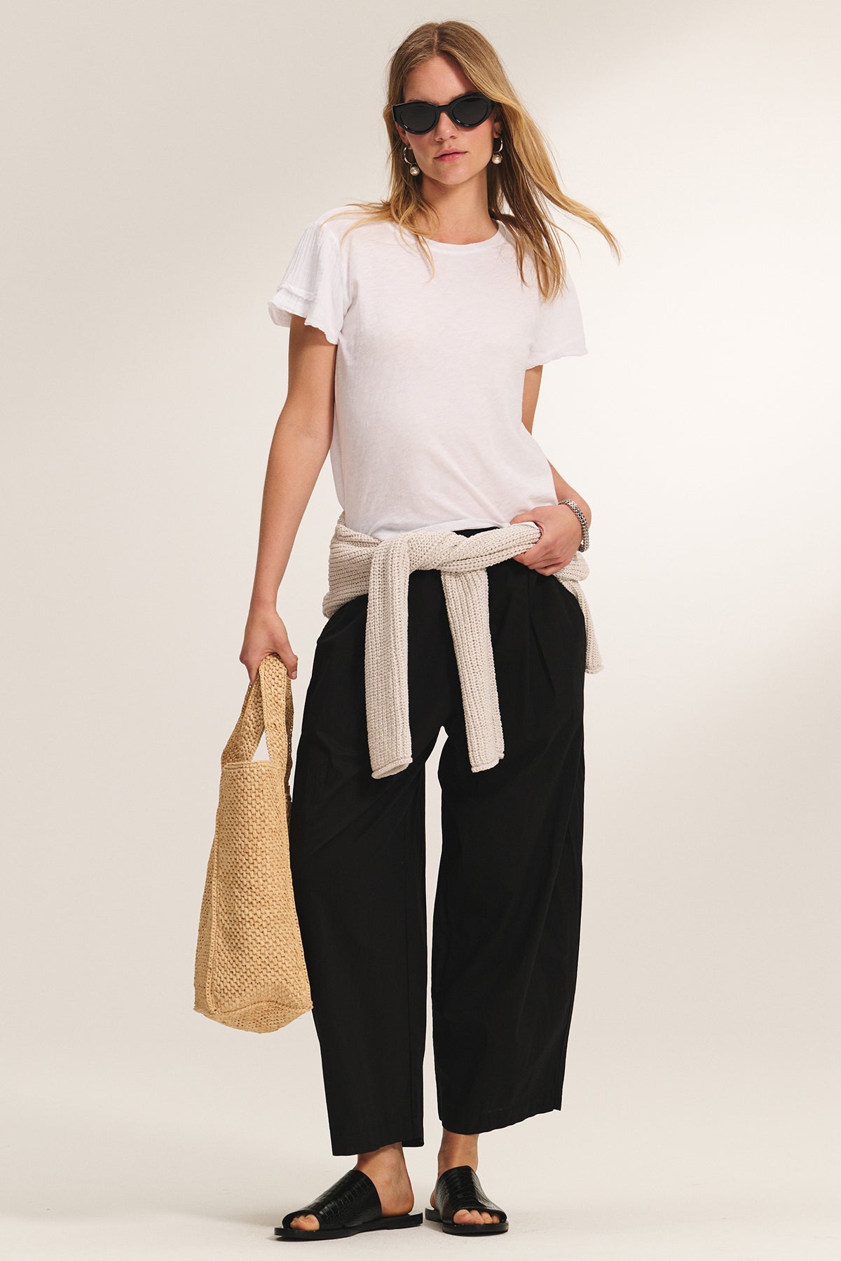 A woman in sunglasses, a white tee, black wide-leg pants, sandals, and the Velvet by Graham & Spencer THALIA CARDIGAN tied at her waist holds a woven bag against a plain background.-39292673523905
