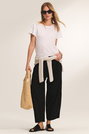 A woman in sunglasses, a white tee, black wide-leg pants, sandals, and the Velvet by Graham & Spencer THALIA CARDIGAN tied at her waist holds a woven bag against a plain background.