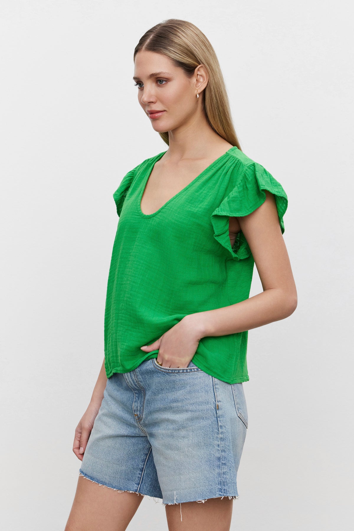   A person poses against a plain background, wearing the REMI RUFFLE SLEEVE TOP by Velvet by Graham & Spencer, a green short-sleeved top with ruffled sleeves made from textured cotton gauze, and blue denim shorts. 