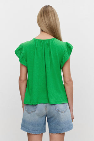 A person with long hair wearing a green REMI RUFFLE SLEEVE TOP by Velvet by Graham & Spencer made of vintage-soft slub knit and blue denim shorts stands facing away, evoking a spring vibe.