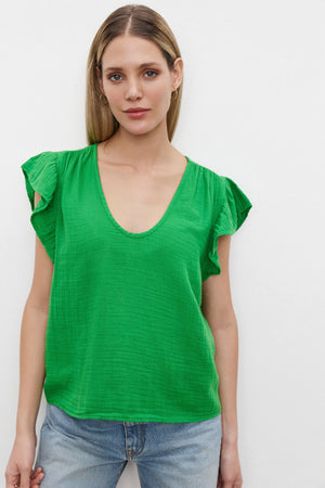 A woman with long blonde hair wearing the REMI RUFFLE SLEEVE TOP by Velvet by Graham & Spencer, a green short-sleeve V-neck blouse made of vintage-soft slub knit, and blue jeans stands against a white background, exuding a spring vibe.