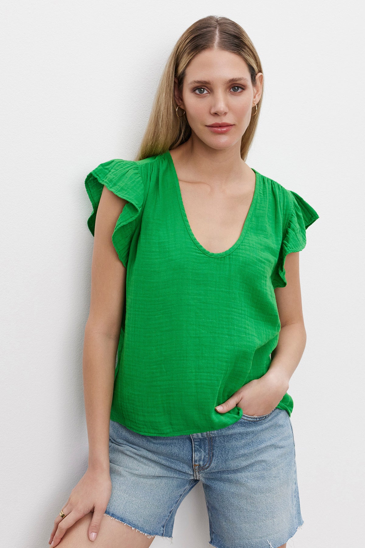 A woman with long blonde hair stands against a white wall, wearing the REMI RUFFLE SLEEVE TOP by Velvet by Graham & Spencer, made of vintage-soft slub knit in green, paired with blue denim shorts. She exudes a spring vibe with one hand in her pocket.-37618565087425