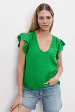 A woman with long blonde hair stands against a white wall, wearing the REMI RUFFLE SLEEVE TOP by Velvet by Graham & Spencer, made of vintage-soft slub knit in green, paired with blue denim shorts. She exudes a spring vibe with one hand in her pocket.