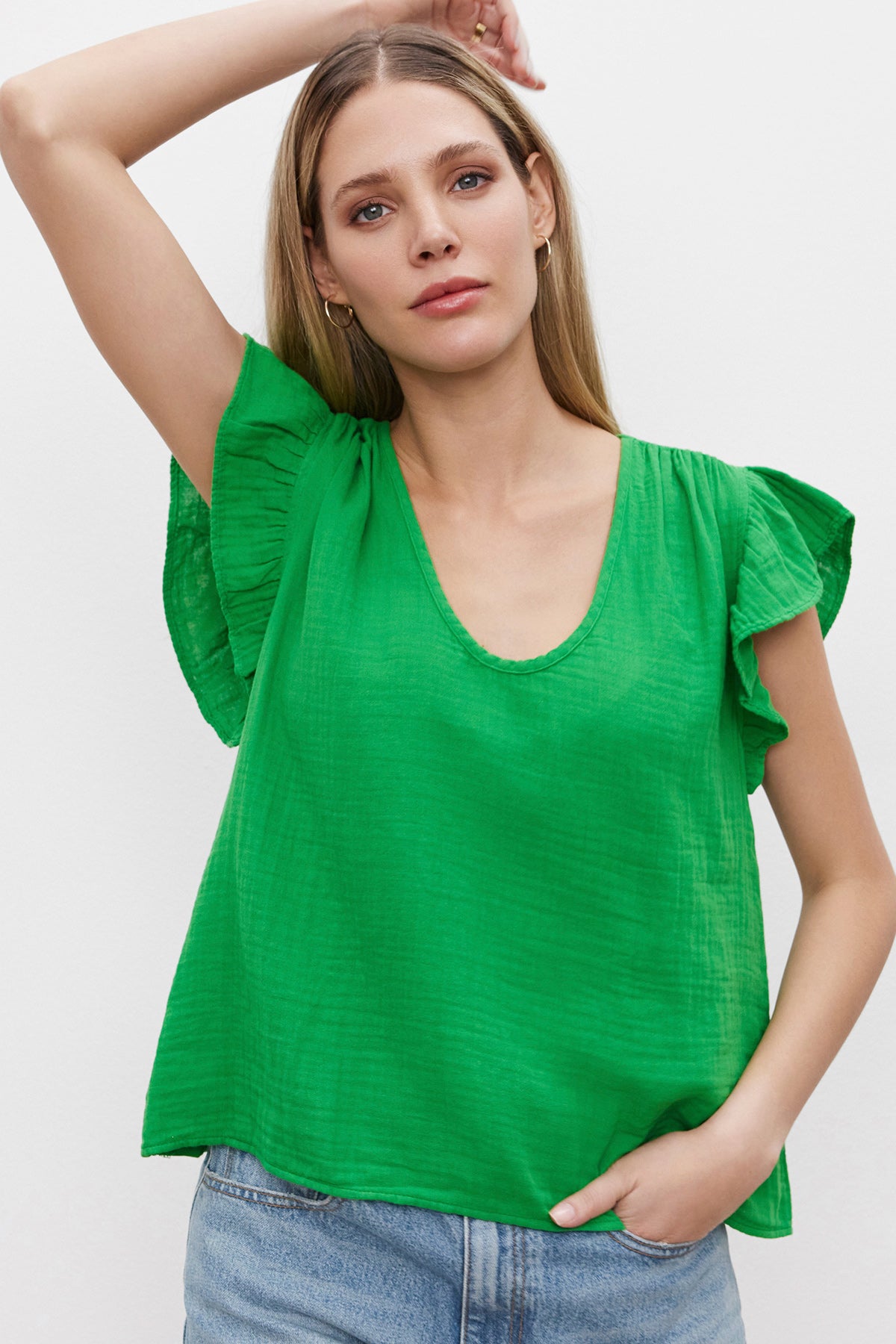   A woman with blonde hair wearing the REMI RUFFLE SLEEVE TOP by Velvet by Graham & Spencer, a green top with ruffled sleeves made from vintage-soft slub knit, and blue jeans stands with one arm raised against a plain white background. 