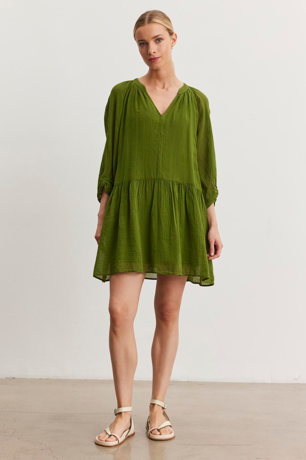   A person stands facing forward, wearing a loose-fitting green V-neck Sloan Dress from Velvet by Graham & Spencer, made from breathable cotton gauze, paired with beige sandals. The background is a plain, light-colored wall. 