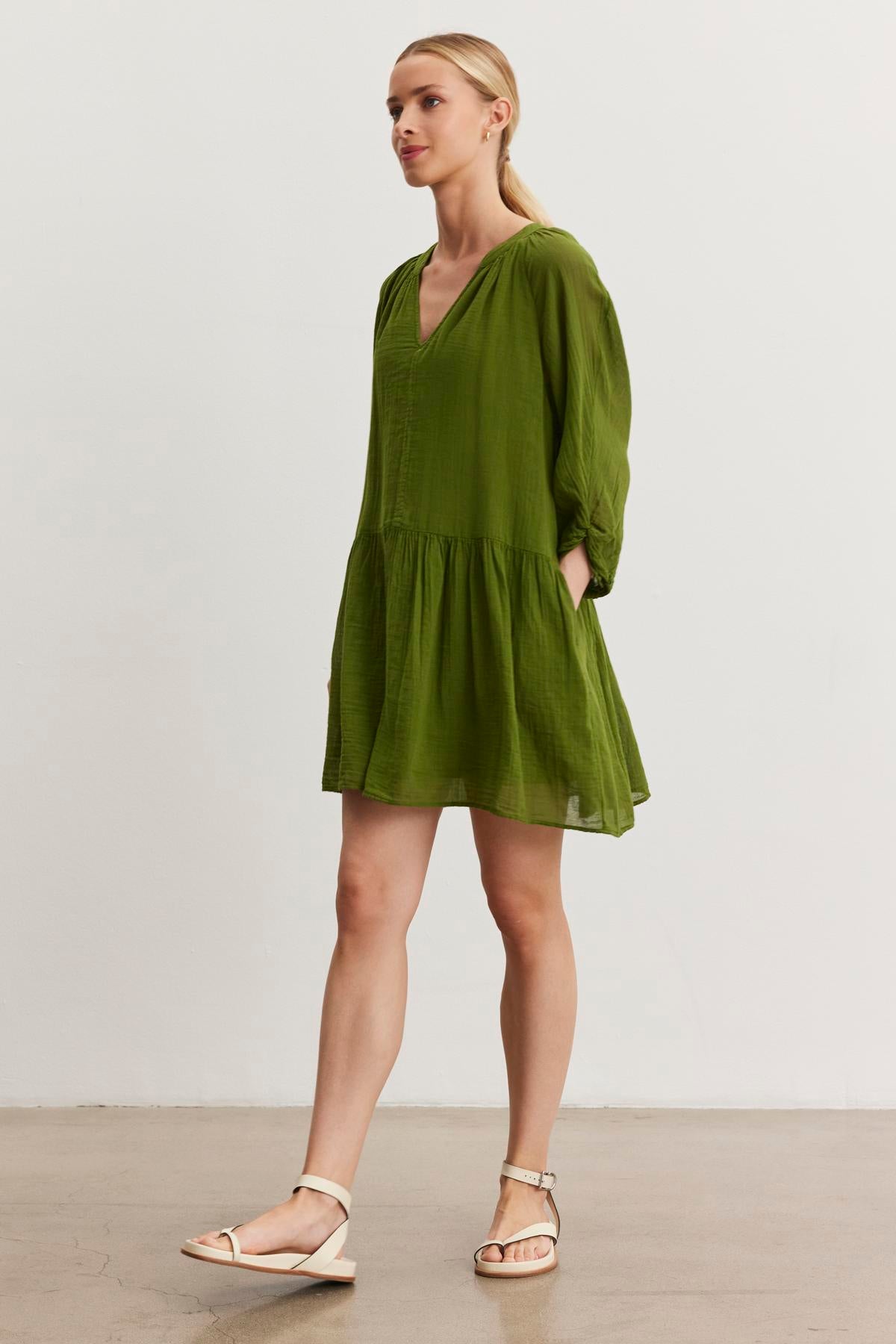   A woman in a green, long-sleeved, knee-length Sloan Dress by Velvet by Graham & Spencer, featuring a V-neckline and white sandals, stands against a plain white background, showcasing the breathable cotton gauze fabric that's perfect for any occasion. 