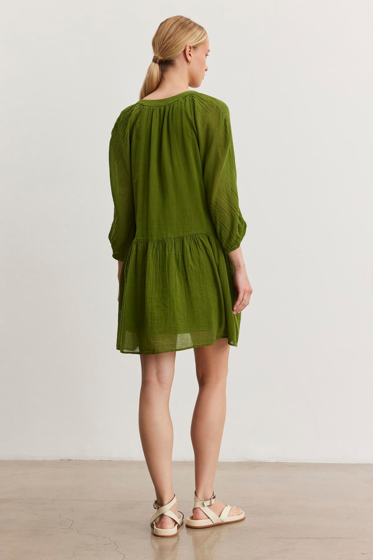   A woman with blonde hair is standing with her back to the camera, wearing the versatile SLOAN DRESS from Velvet by Graham & Spencer, a green long-sleeved dress made of breathable cotton gauze. She is also wearing white sandals. 