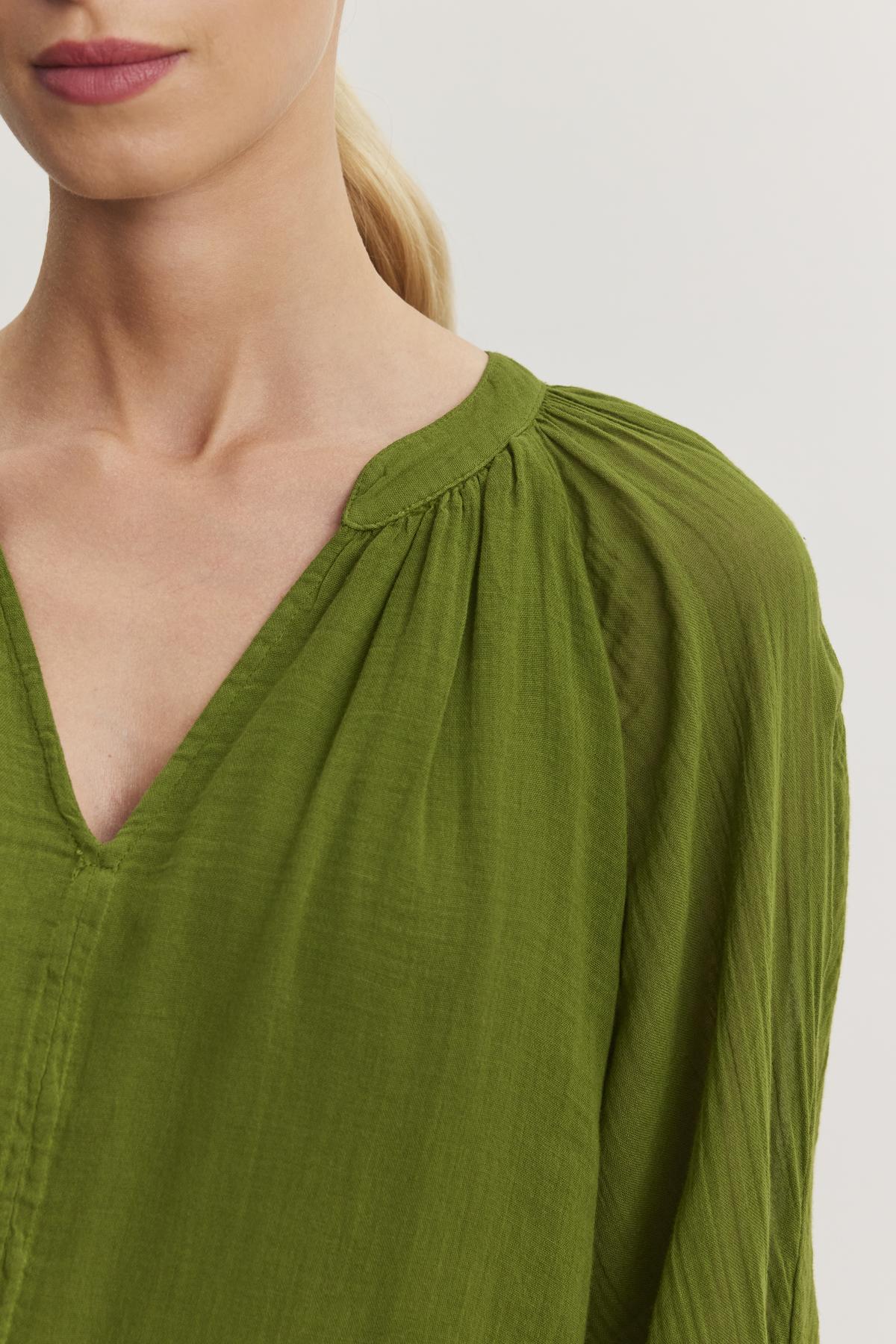   A person with light skin and blonde hair in a ponytail wears a green, v-neck, long-sleeve blouse with gathered fabric details at the shoulders from Velvet by Graham & Spencer, showcasing a versatile piece reminiscent of the Sloan Dress. 