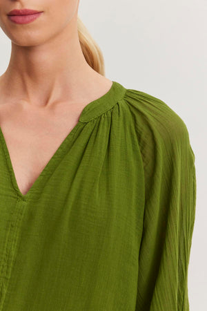 A person with light skin and blonde hair in a ponytail wears a green, v-neck, long-sleeve blouse with gathered fabric details at the shoulders from Velvet by Graham & Spencer, showcasing a versatile piece reminiscent of the Sloan Dress.