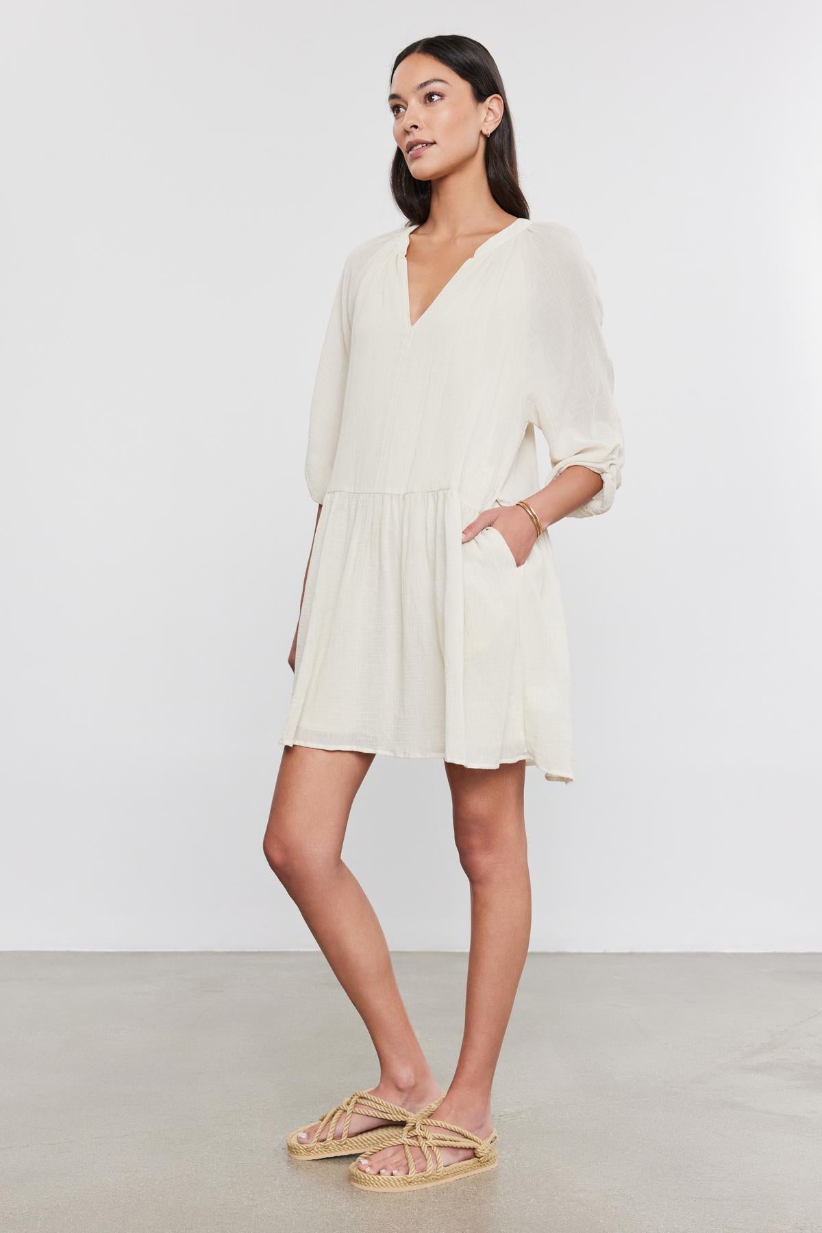   A woman in a white Velvet by Graham & Spencer Sloan Dress with pleat detailing and beige sandals stands against a gray background, looking to the side with her hands slightly behind her. 